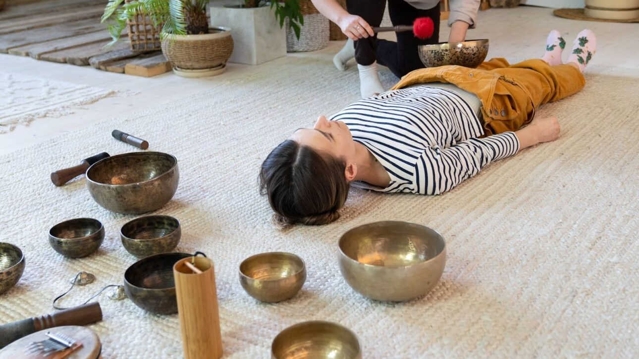 A facilitator leads a woman to an Asian-inspired sound bath using bronze singing bowls and a gong.