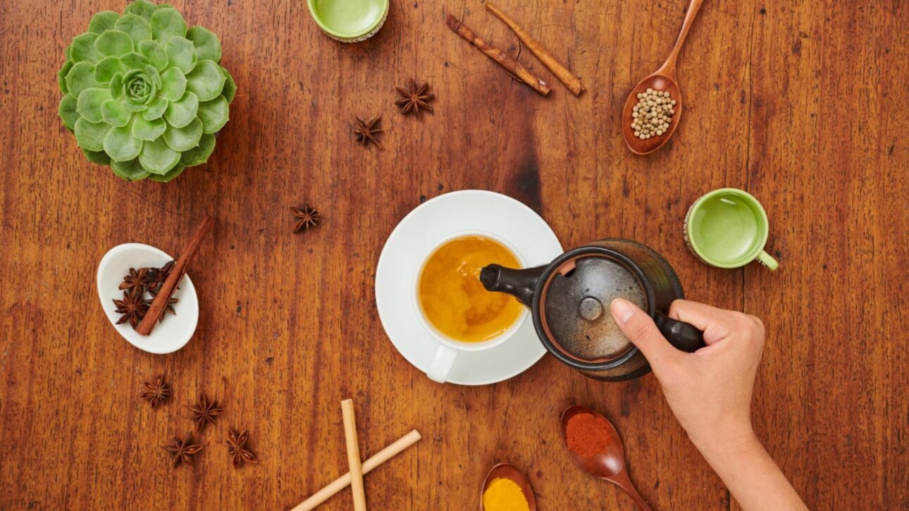 Woman pouring tea into a cup next to herbs, spices, and plants.