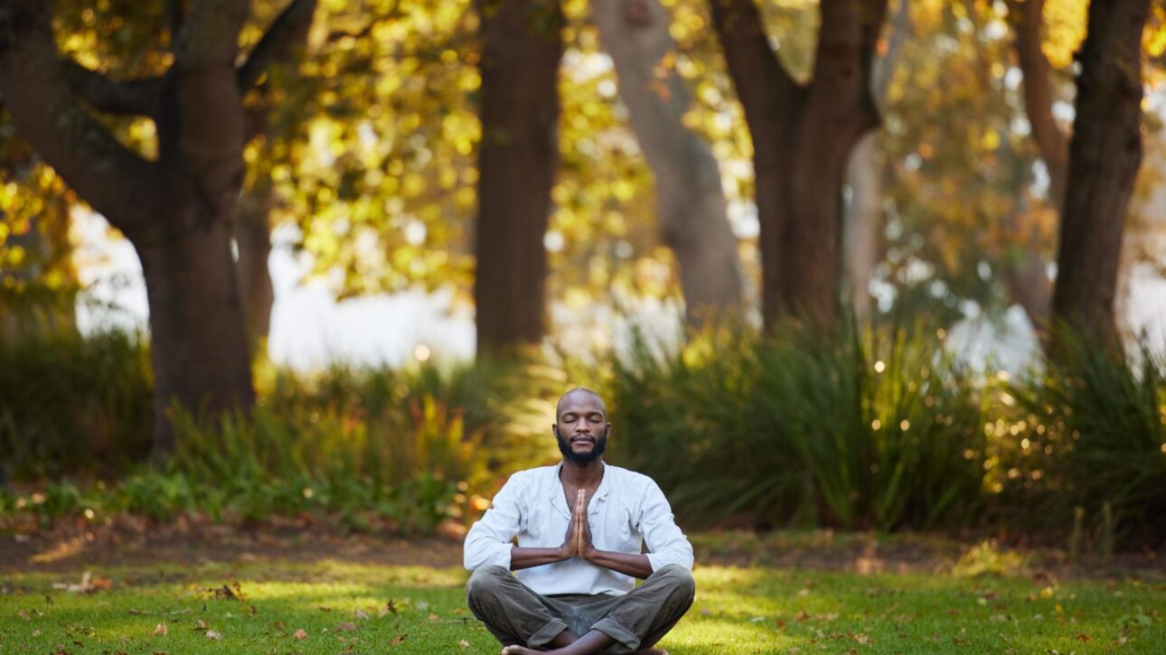 Full-body shot of a young man meditating outdoors in a park.